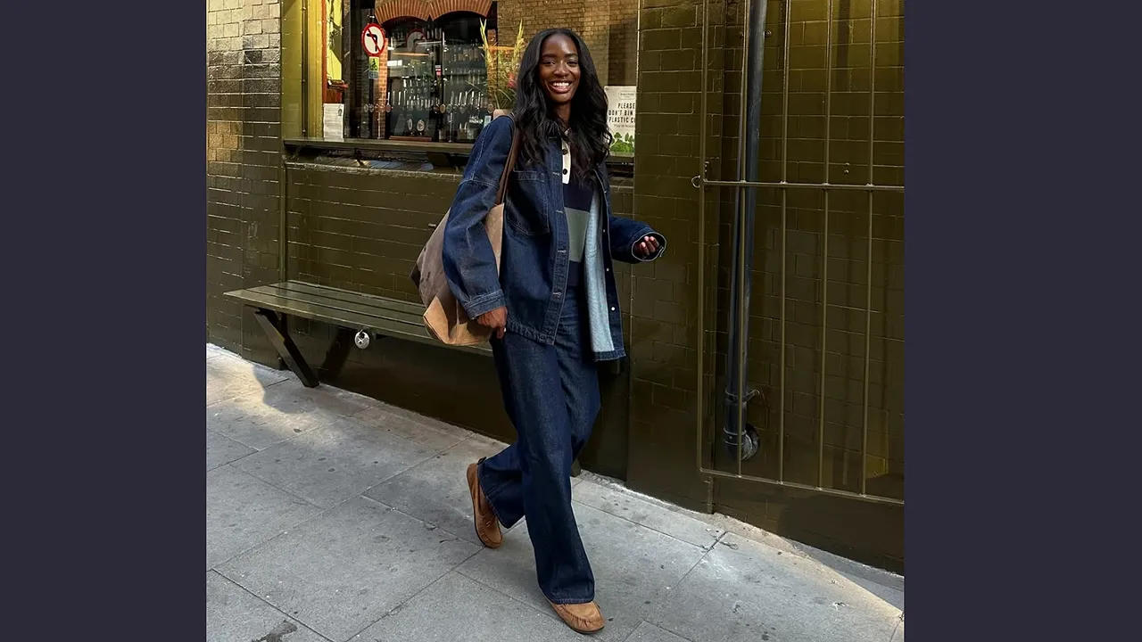 Woman wearing a dark blue jacket and dark jeans with brown shoes beside a green storefront