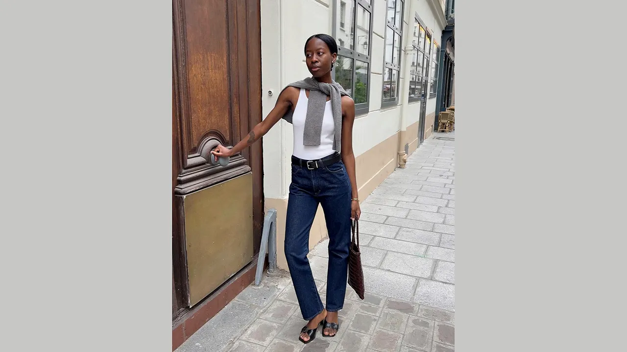 Woman wearing dark blue jeans with a white sleeveless top, a sweater over her shoulders, and heeled shoes